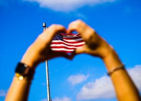 manos en forma de corazón con la bandera norteamericana a lo lejos entre ellas