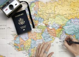 Persona marcando destinos en un mapa de papel, donde se ven también un pasaporte norteamericano y una cámara fotográfica