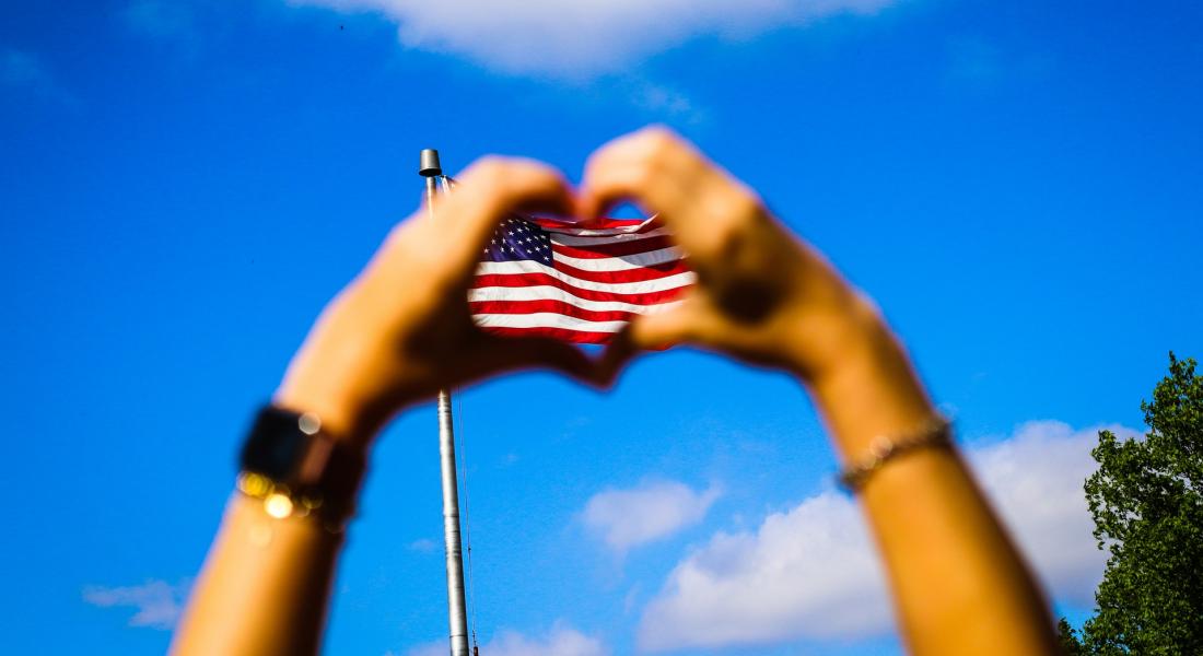 manos en forma de corazón con la bandera norteamericana a lo lejos entre ellas
